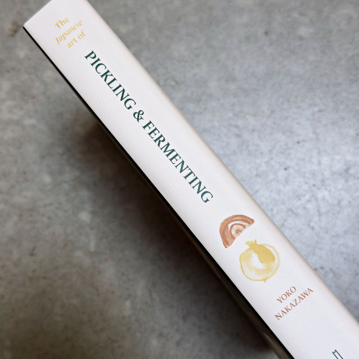 A close-up diagonal shot of a book spine titled 'The Japanese art of PICKLING & FERMENTING' by Yoko Nakazawa, featuring a cream-colored cover with dark green text and small yellow and brown illustrations, set against a textured grey background.