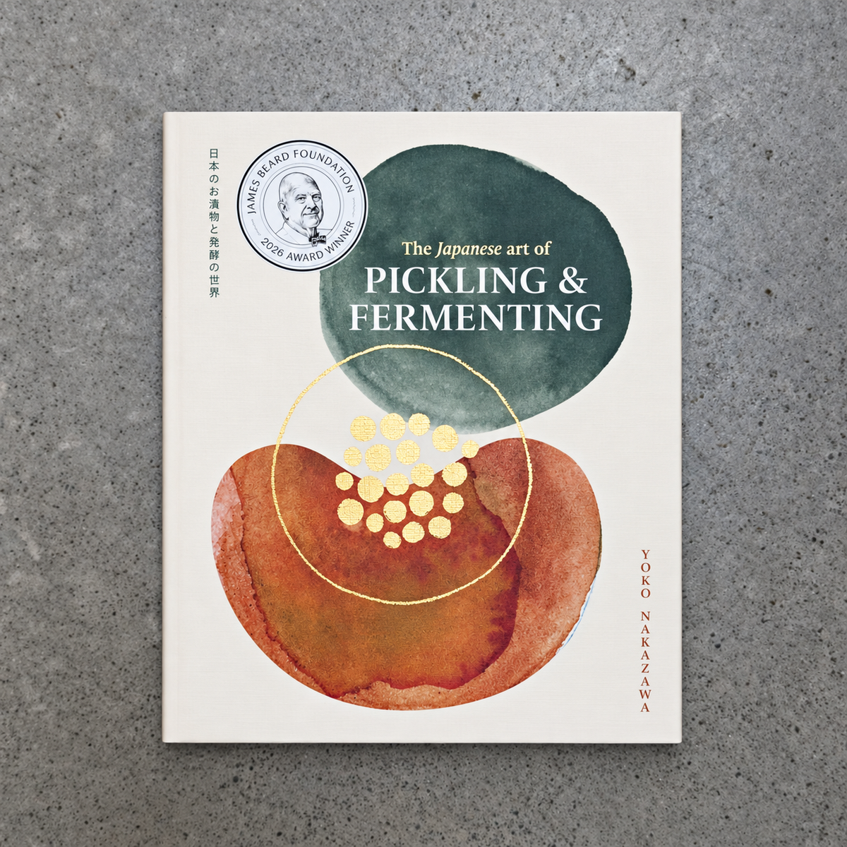A book titled "The Japanese art of Pickling & Fermenting" by Tokyo Nakazawa lies flat on a textured grey concrete surface. The cover features an off-white background with abstract watercolour shapes in dark green and terracotta orange, accented by gold circles and text.