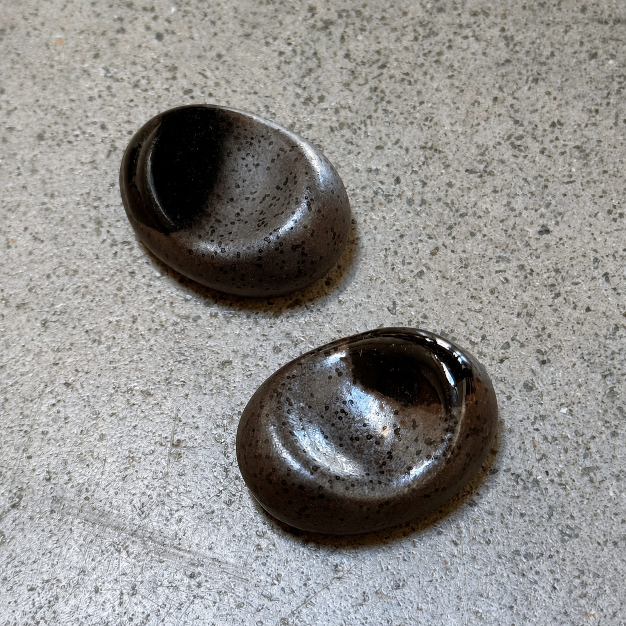 Two oval Japanese ceramic chopstick rests with a dark brown and black speckled glaze, resting on a textured grey surface.