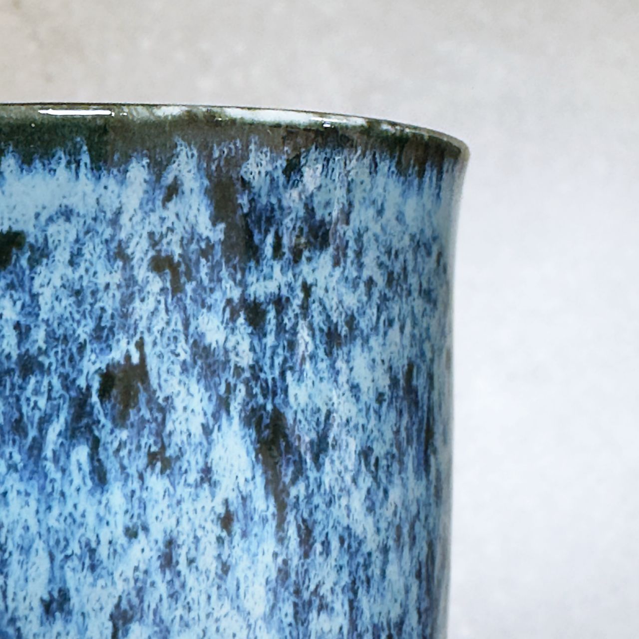 Close-up of a ceramic cup with a flowing blue and dark blue speckling, shown against a light neutral background.