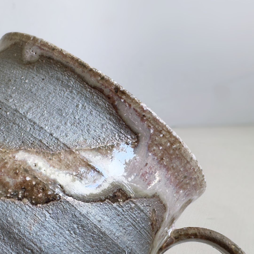 Close-up of the mug rim showing textured dark clay and softly pooled pale glaze on a pale grey background.