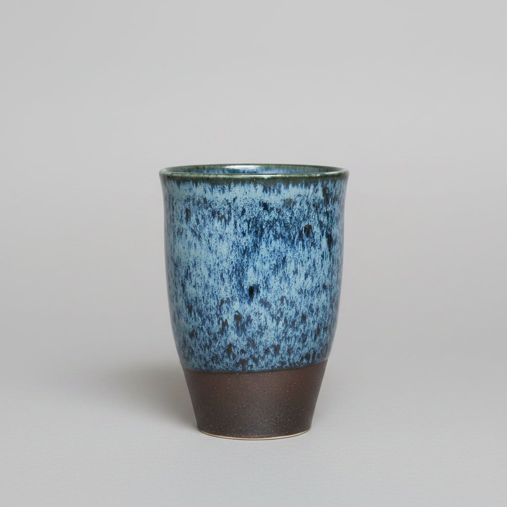 A handmade ceramic cup with a textured blue and bark blue speckled glaze on the upper half and a dark brown unglazed base.