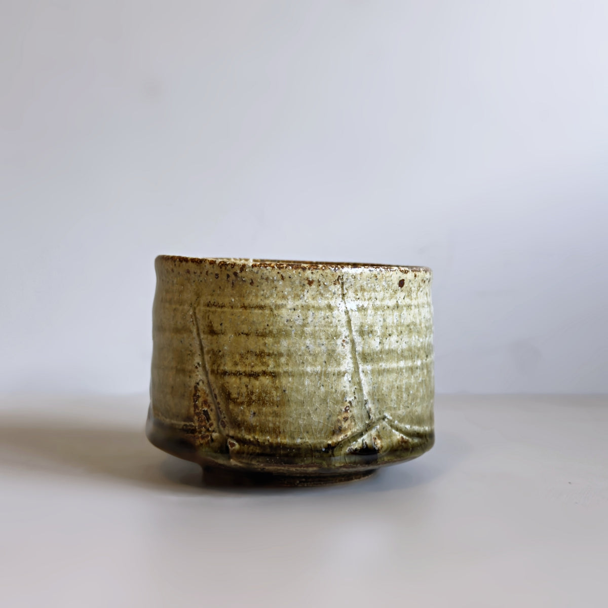 A handmade Japanese ceramic matcha bowl with a textured, rustic glaze in muted olive green and brown tones, set against a plain light grey background.
