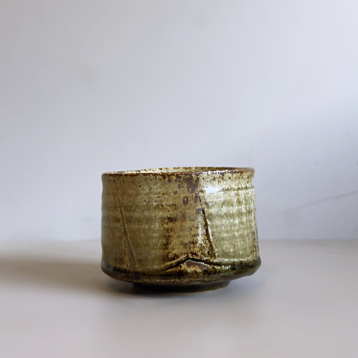 A handmade Japanese ceramic matcha bowl with a textured, rustic glaze in muted olive green and brown tones, set against a plain light grey background.