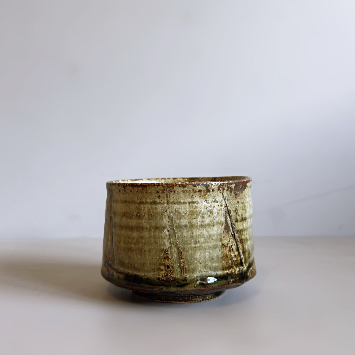 A front view of a handmade Japanese ceramic matcha bowl with a textured, rustic glaze in muted olive green and brown tones, set against a plain light grey background.