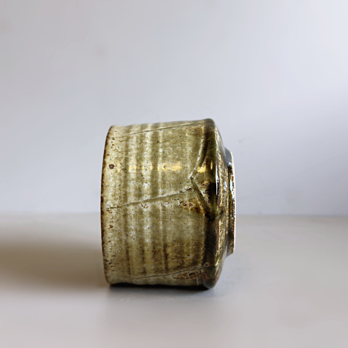 A handmade Japanese ceramic matcha bowl with a textured, ribbed exterior and a glossy, metallic olive-green glaze, standing on a neutral light grey surface against a white background.