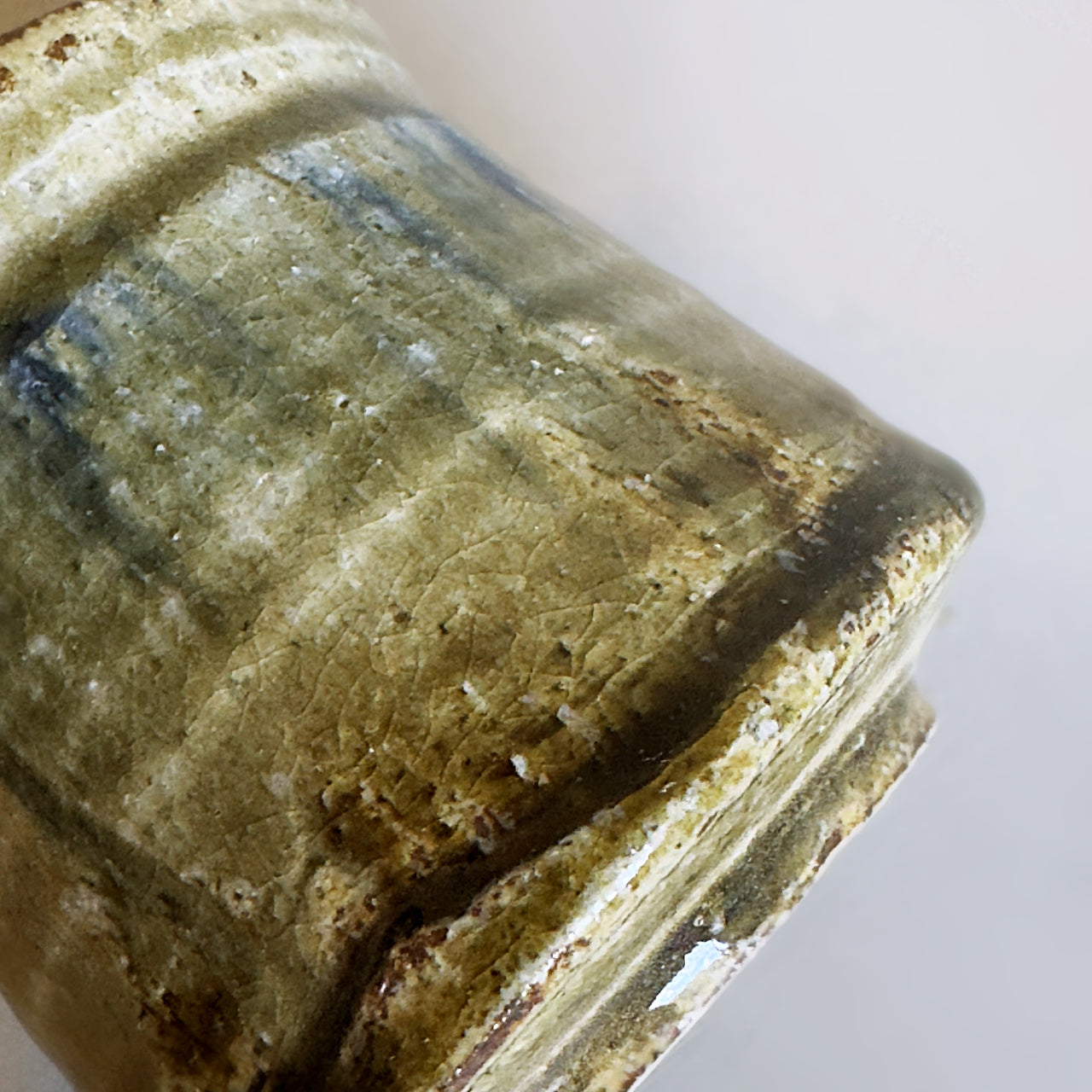 Close-up of a handmade ceramic vessel showing its textured, rustic glaze with earthy green, brown, and beige tones.