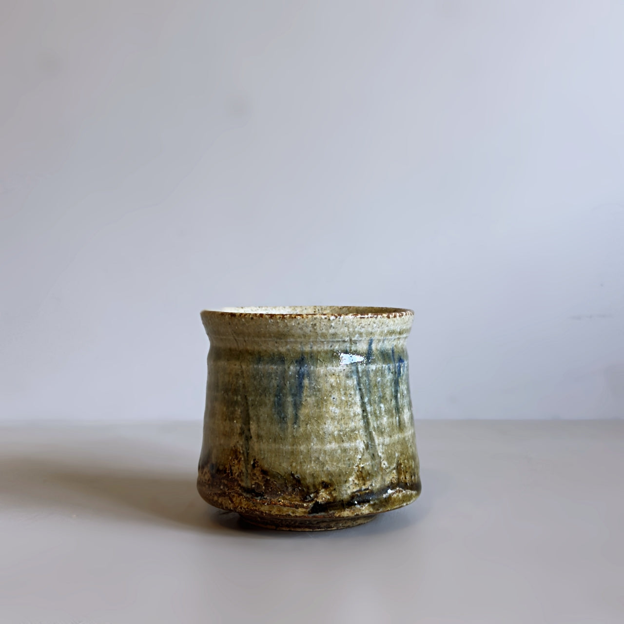 A handmade Japanese ceramic teacup with a rustic, tapered shape, featuring a textured glaze with vertical blue-green and brown drips over a beige base, set against a plain light grey background.
