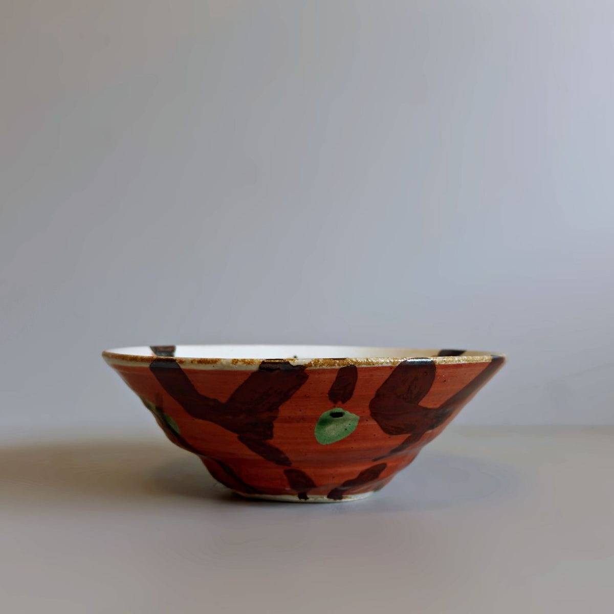 A handmade Japanese ceramic bowl with a red glaze exterior decorated with dark brown brushstrokes and a small green accent, set against a plain light grey background.