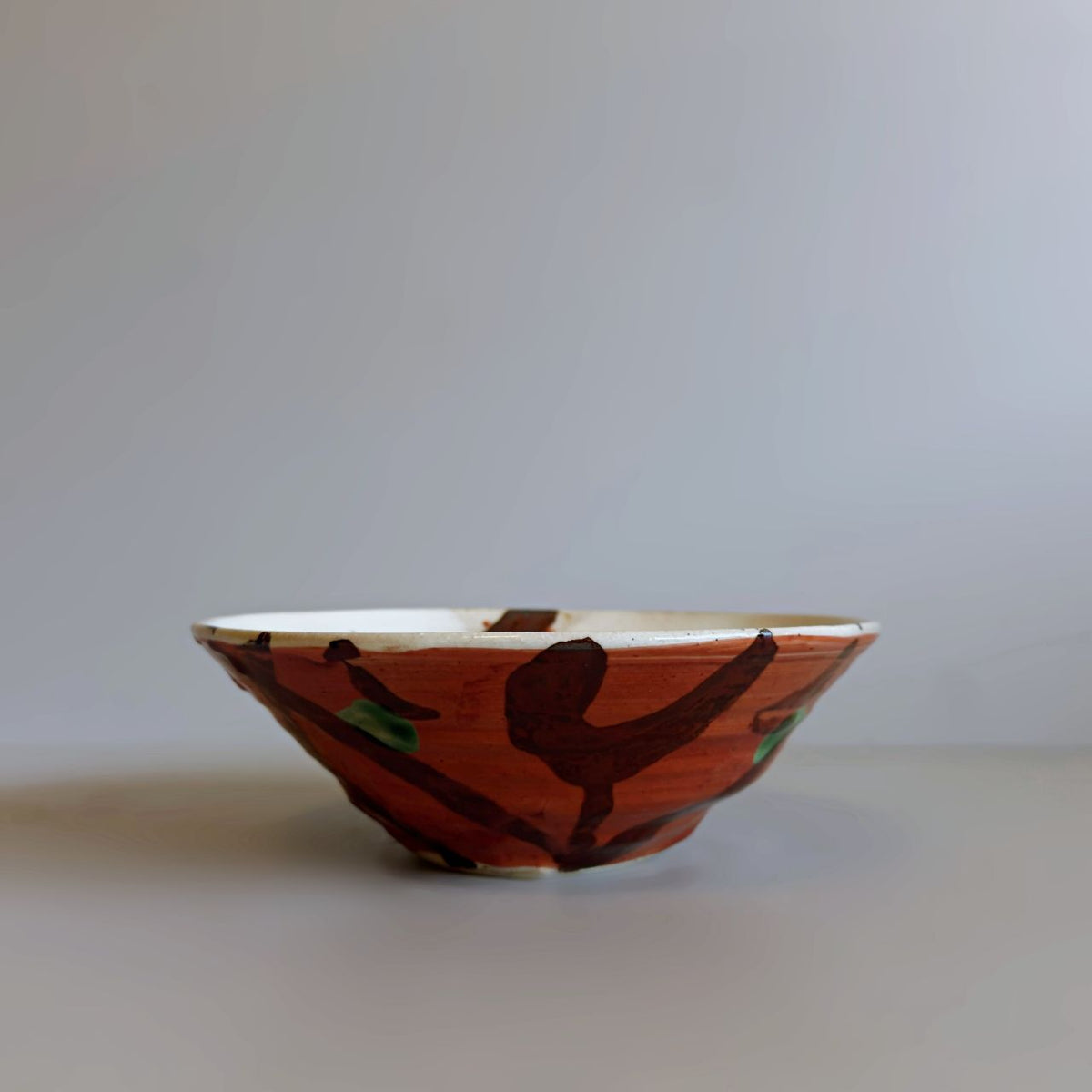 A handmade Japanese ceramic bowl with a red glaze decorated with abstract hand-painted brown and green patterns, set against a neutral grey background.