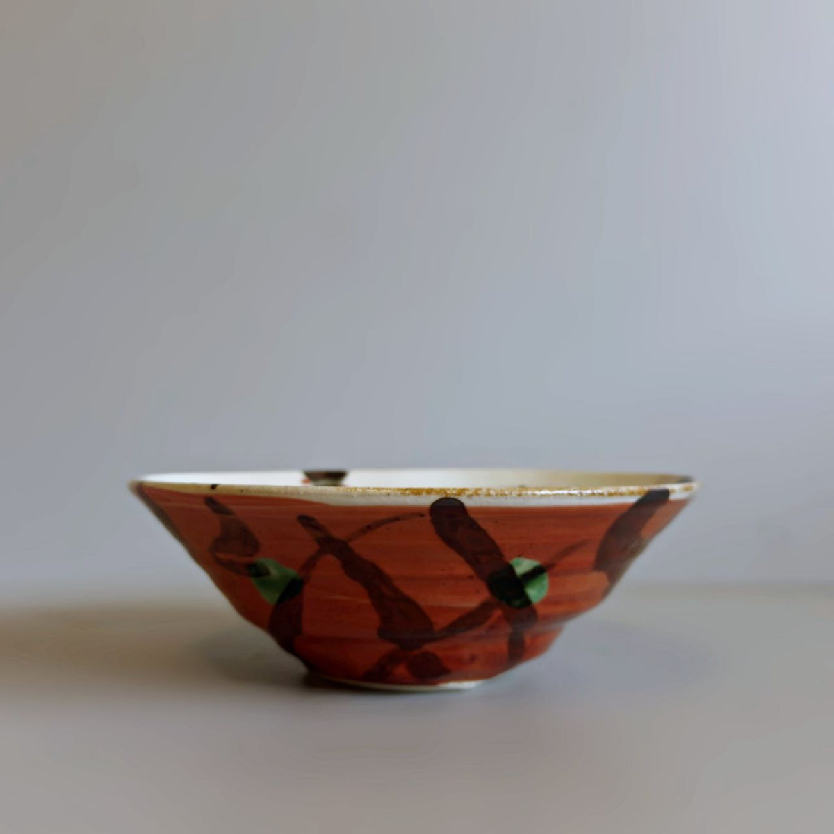 A handmade Japanese ceramic bowl with an earthy red glaze, decorated with dark brown brushstrokes and small green accents, set against a plain light grey background.