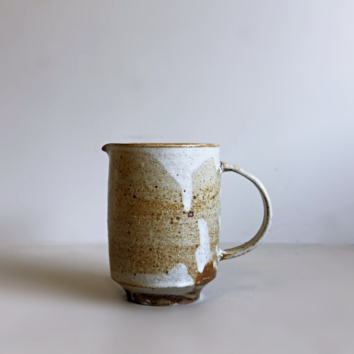 A handmade ceramic jug with a rustic cream and brown speckled glaze, shown from the front against a pale grey background.
