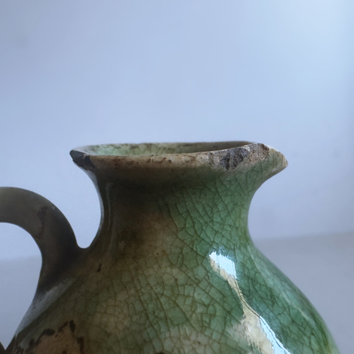 Close-up of a vintage ceramic vase with a green glaze and crackle pattern, featuring a handle and a flared rim against a light grey background.