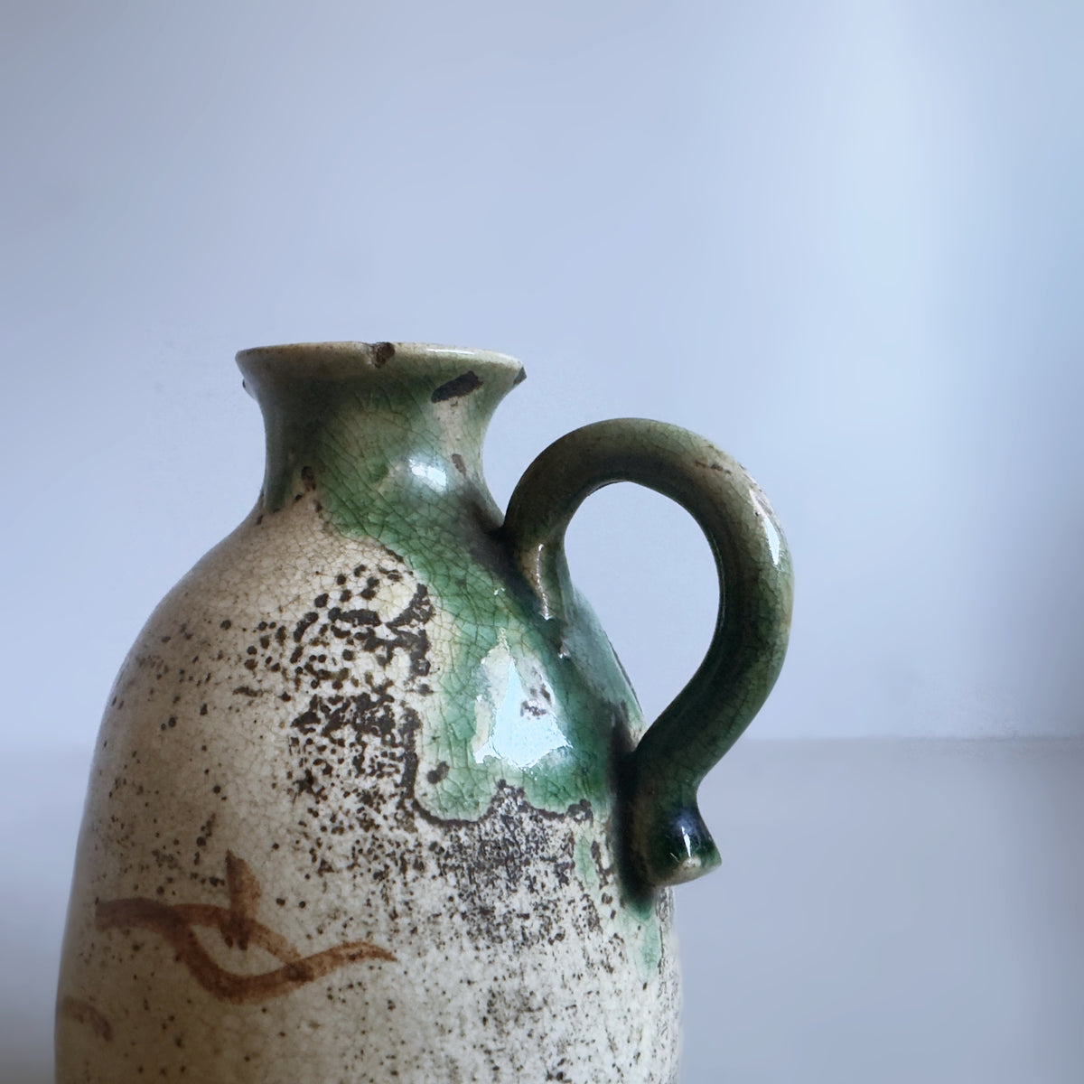 A handmade ceramic pitcher with a rounded body, a curved handle, and a small spout. It features a speckled cream glaze with dark brown spots and a glossy green glaze dripping over the top and along the handle.