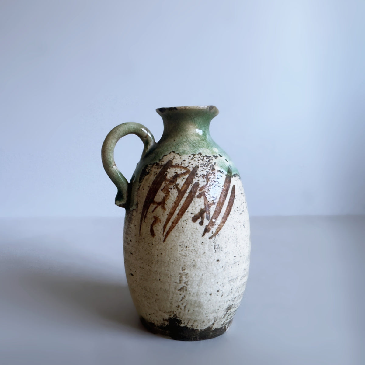 A vintage handmade ceramic jug with a rounded body, a side handle, and a narrow neck, featuring a speckled off-white glaze with brown brushstrokes and a green glaze on the neck and handle, set against a plain light grey background.