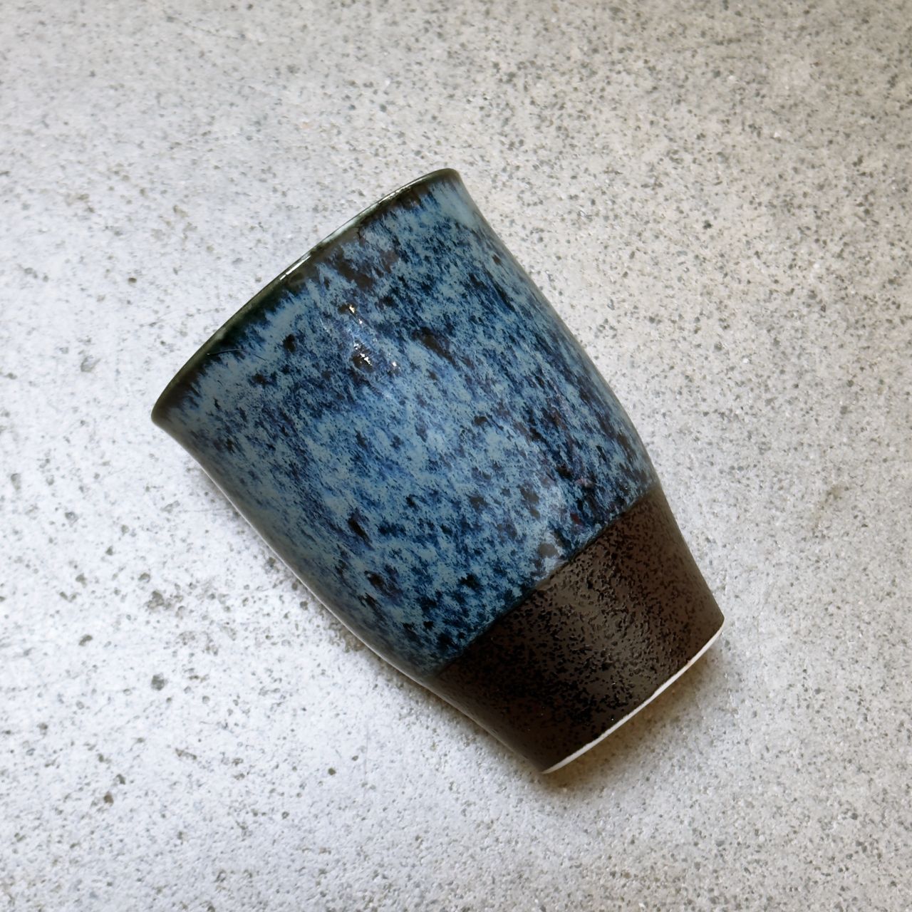 A handmade ceramic cup with a textured blue speckled glaze on the upper half and a dark brown glaze band at the base, resting on a  light grey concrete surface.