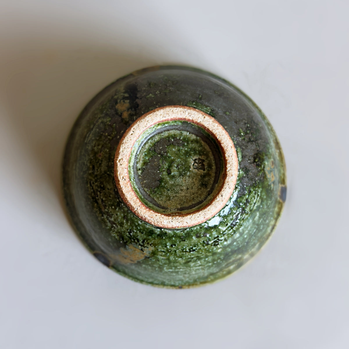 Top-down view of a bottom of a Japanese ceramic bowl with a textured green oribe glaze and a contrasting beige clay rim, photographed on a neutral light grey background.