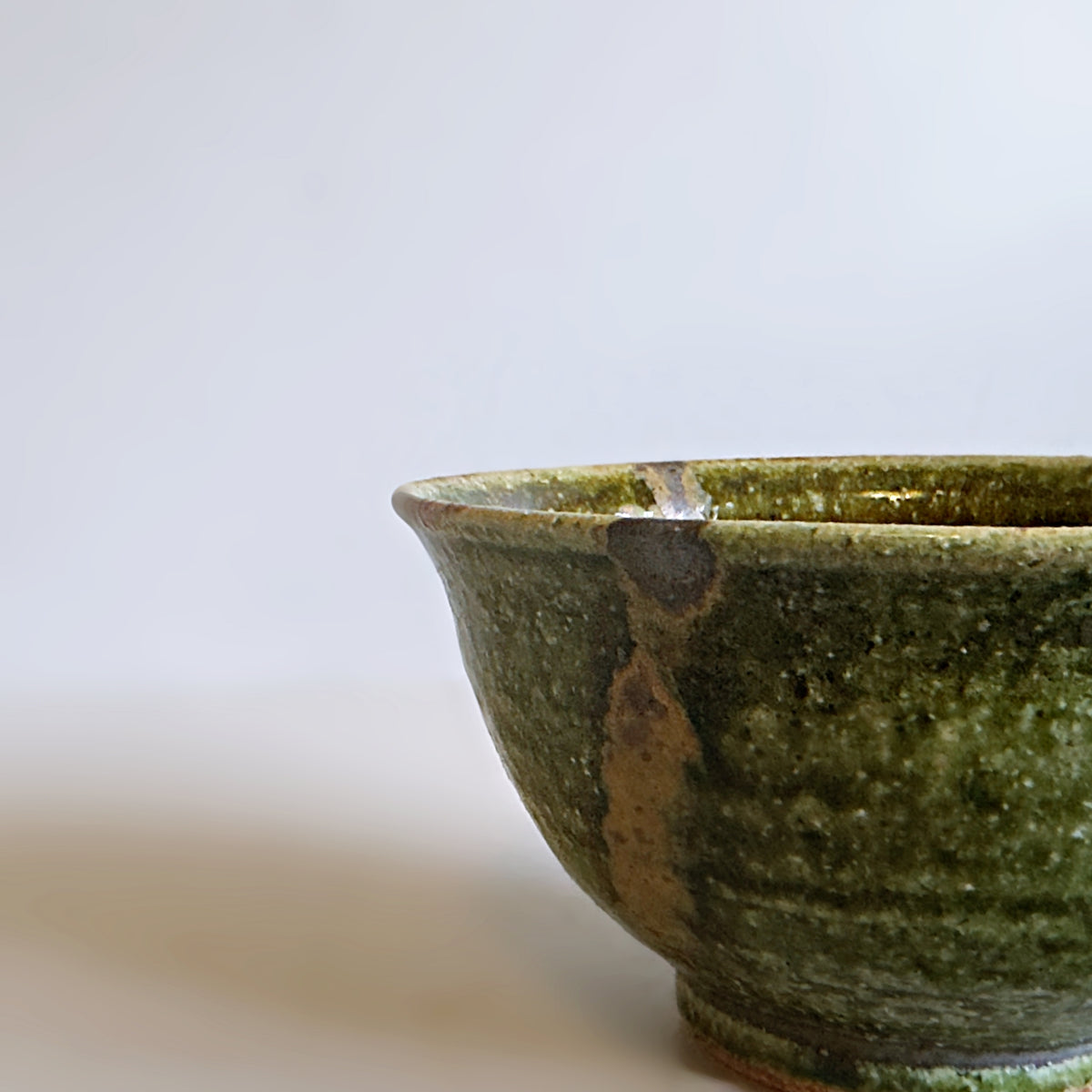 A close-up of a handmade Japanese ceramic bowl with a textured green oribe glaze and a natural brown crack, set against a plain light grey background.