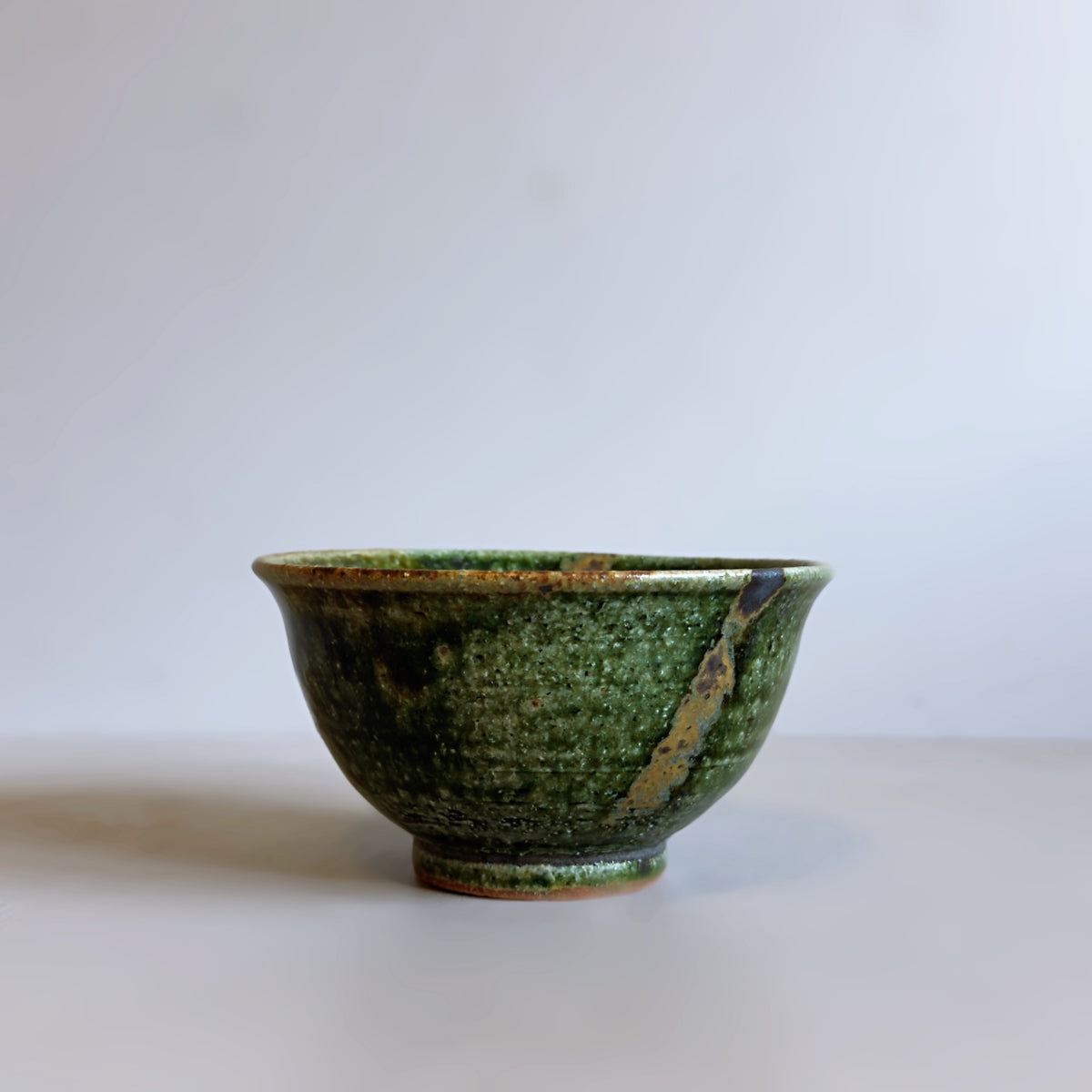A Japanese handmade ceramic rice bowl with a textured green oribe glaze and natural brown toned crackle patterns, photographed on a neutral light grey background.