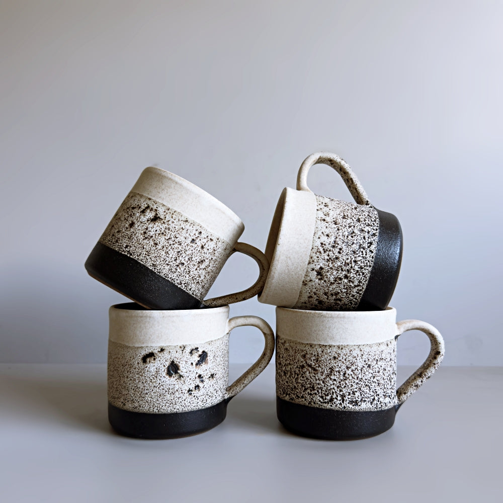 Four Japanese Mino ware ceramic mugs with speckled design and black bases on a light grey background.