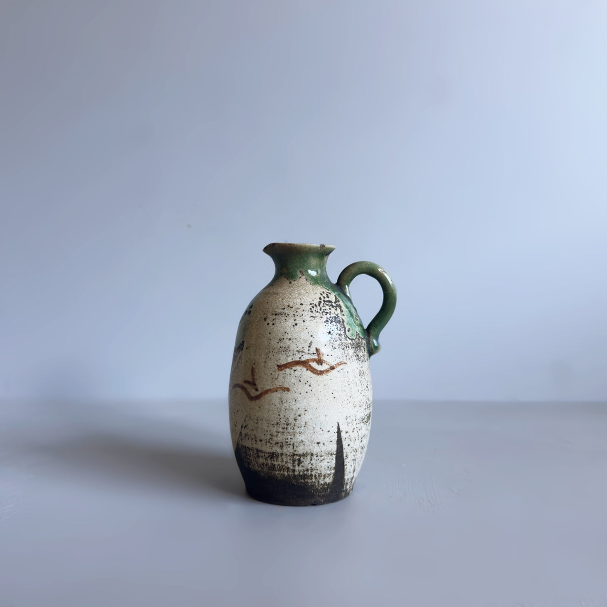 A vintage small handmade ceramic vase with a rounded body, a side handle, and a short spout, featuring a distressed off-white glaze with green and brown accents, set against a plain light grey background.