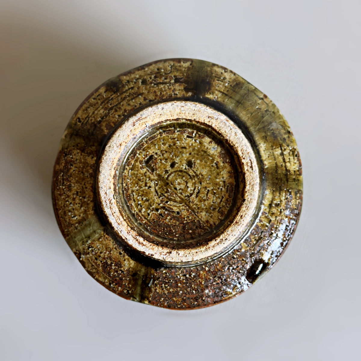 A top-down view of a handmade ceramic Japanese matcha bowl with a rustic, glazed finish in earthy brown, green, and gold tones, set against a plain light grey background.