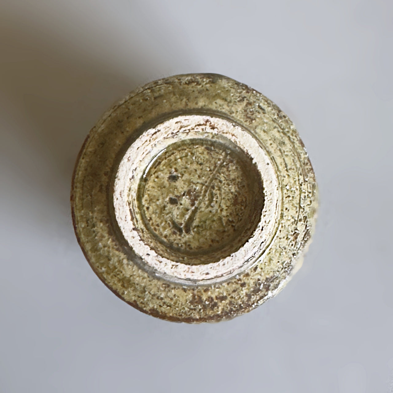 Top-down view of a handmade Japanese ceramic tea cup base with a textured, speckled glaze in earthy green, brown, and cream tones on a neutral light grey background.
