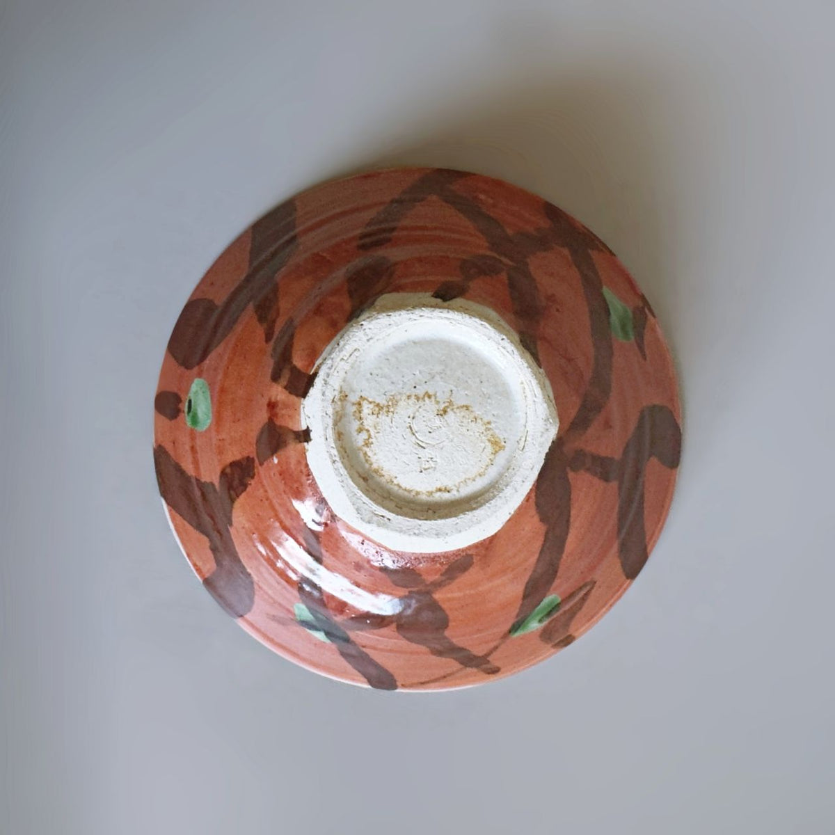 A Japanese handmade ceramic bowl viewed from directly above, featuring an earthy red exterior with dark brown abstract brushstrokes and small green accents, and a white glazed interior base.