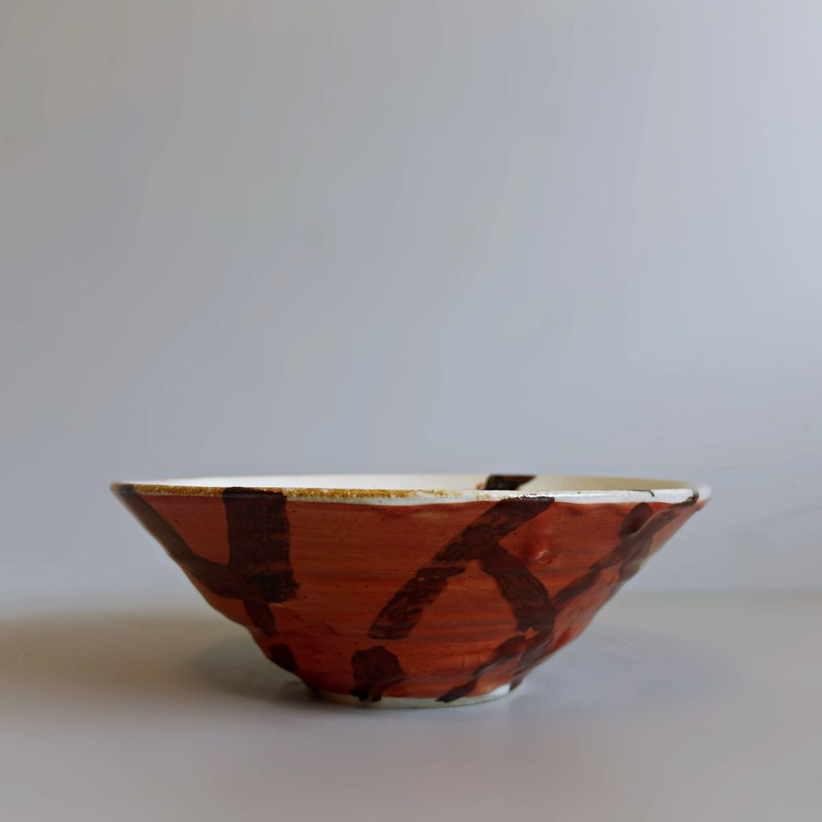 A handmade Japanese ceramic bowl with a red glaze and dark brown abstract brushstroke patterns, set against a plain light grey background.