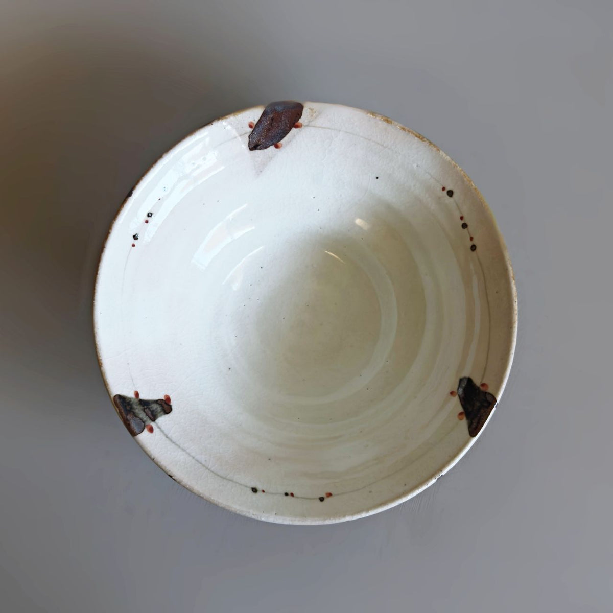 An interior of Japanese off-white interior ceramic bowl with three dark brown spots and small red speckles around the rim, photographed from a top-down perspective on a neutral grey background.