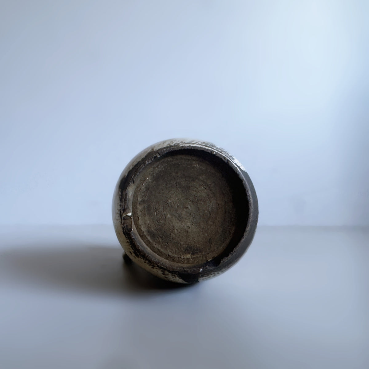 A close-up shot of the textured, circular bottom base of a rustic ceramic vase, showing a dark brown glazed finish against a neutral light grey background.