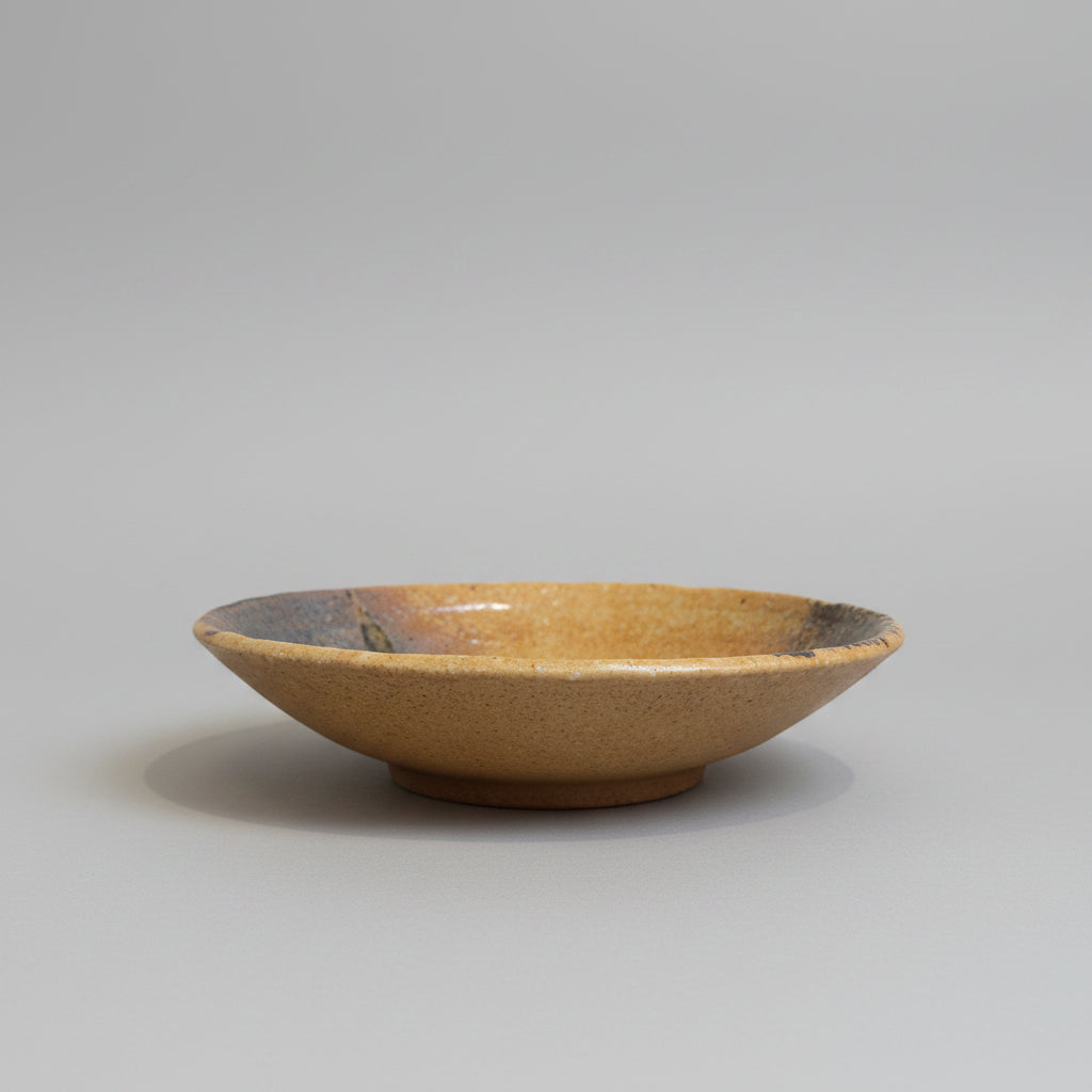 A handmade  Japanese ceramic shallow bowl with a speckled brown exterior and a dark grey glazed interior, sitting on a grey background.