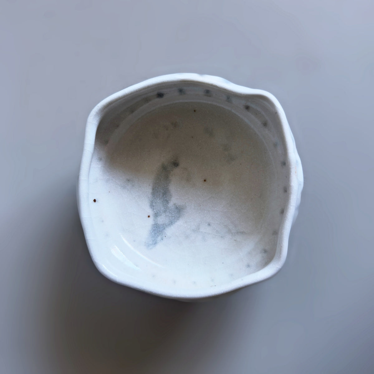 A Japanese handmade ceramic bowl with a scalloped rim, featuring a white speckled glaze and subtle grey speckling details inside, photographed from a top-down perspective on a neutral grey background.